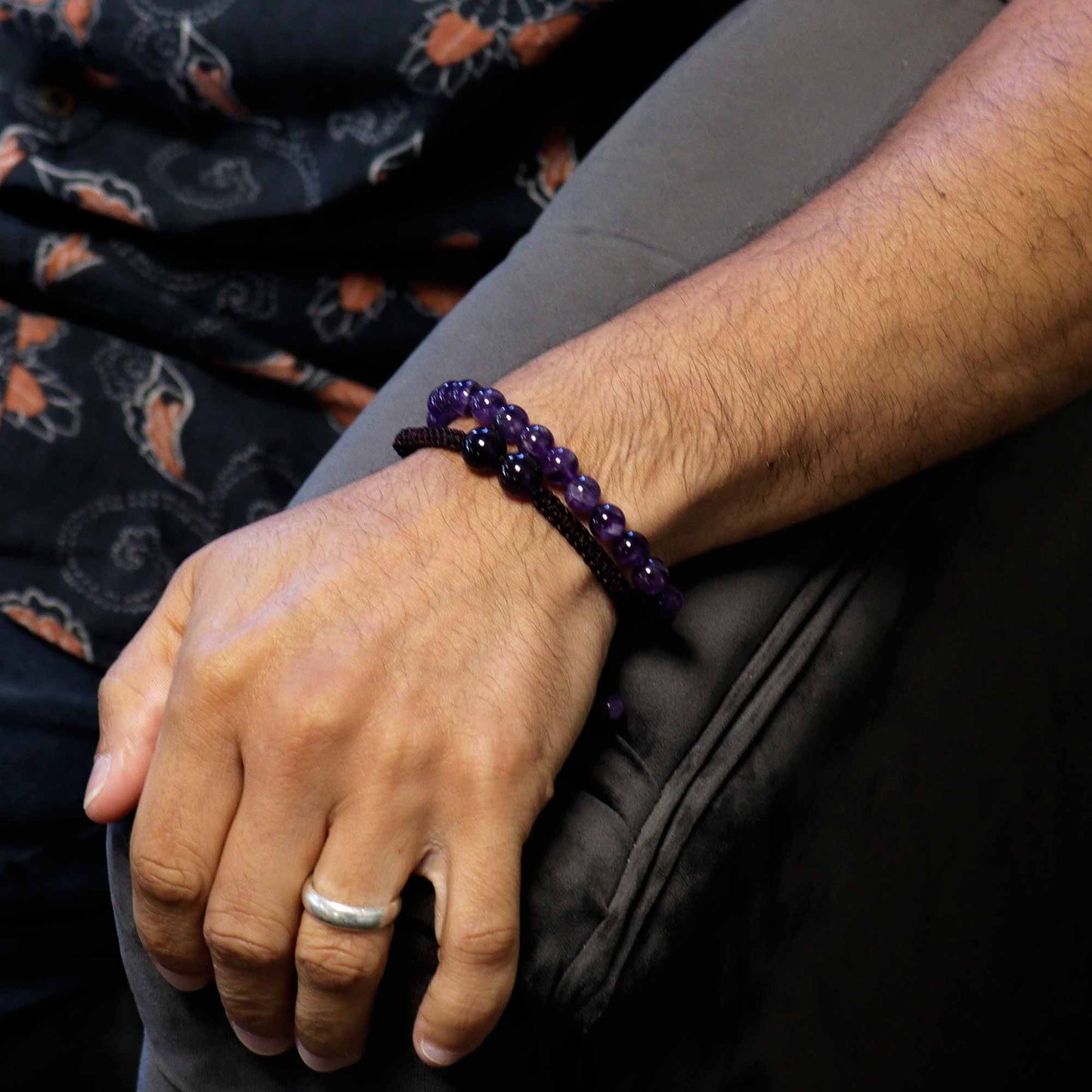 Hand wearing a purple beaded bracelet with a ring, against a dark fabric background