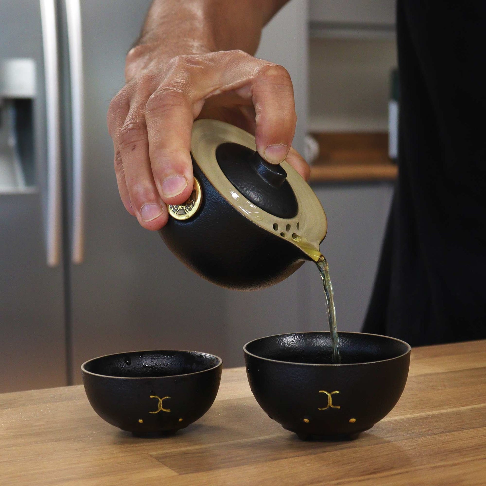 Person pouring tea from a black teapot into two black cups on a wooden surface.